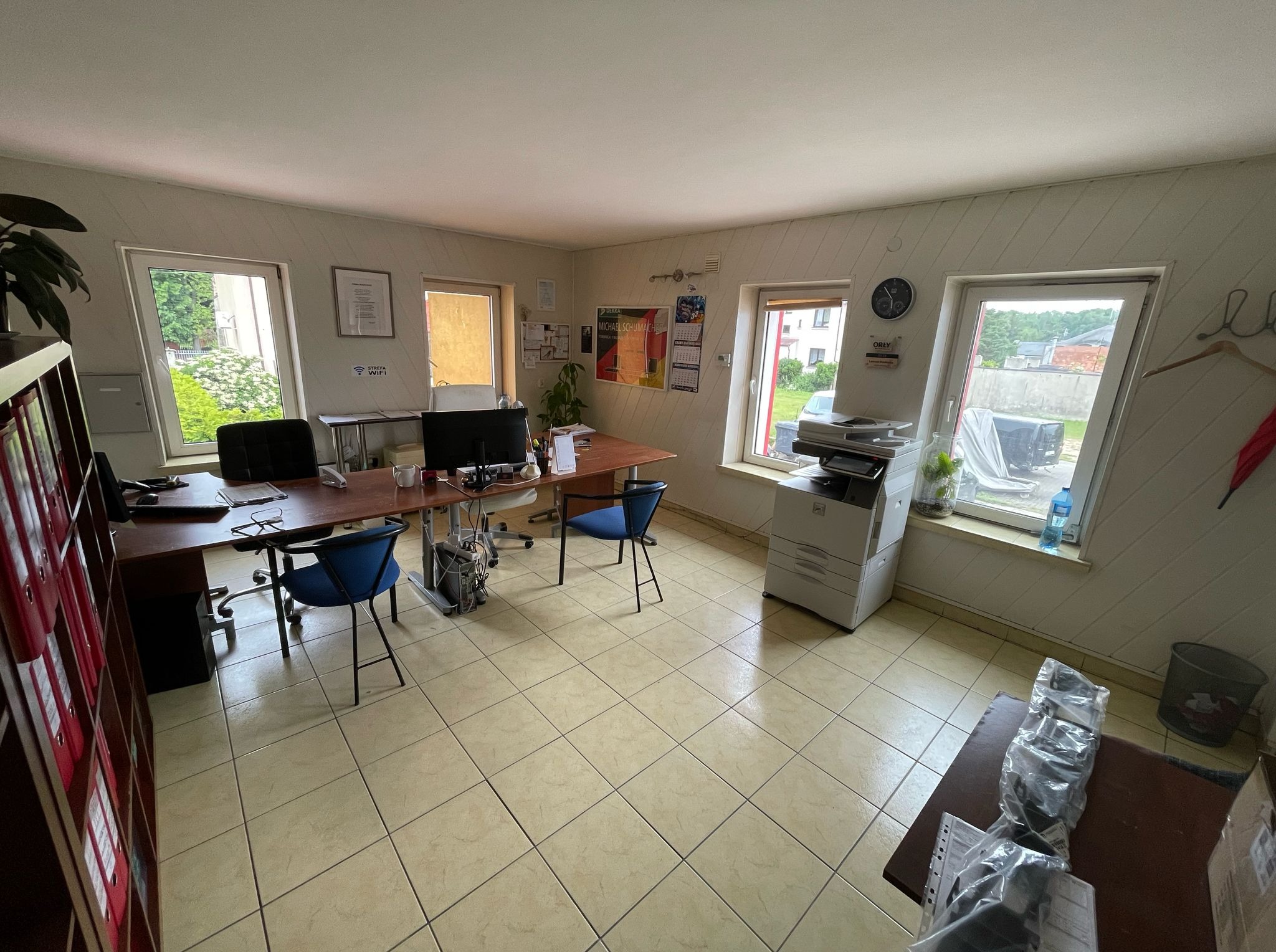 Jasne biuro warsztatu samochodowego z biurkami, krzesłami, komputerami i drukarką. Na ścianach plakaty i dyplomy. Widok na zieleń przez okna.