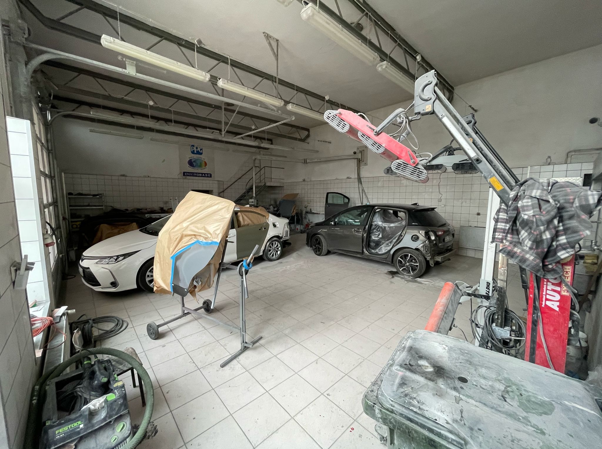 Warsztat: biały samochód z osłoniętą karoserią, szary pojazd w trakcie naprawy, lampa grzewcza i narzędzia blacharskie w jasnym pomieszczeniu z białą glazurą.