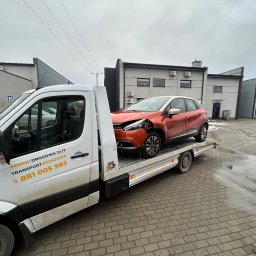 ML Autogroup Mikołaj Lachiewicz - Pomarańczowy uszkodzony samochód na lawecie pomocy drogowej z numerem 881 005 983 na tle szarego budynku w pochmurny dzień.