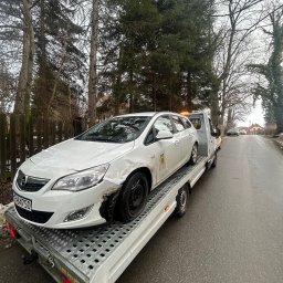 ML Autogroup Mikołaj Lachiewicz - Biała Opel Astra z uszkodzonym przodem na lawecie na tle drzew i drogi. Widoczne tablice rejestracyjne i światła ostrzegawcze. Laweta na drodze w pochmurny dzień.