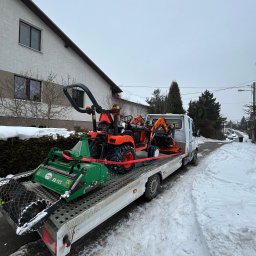 ML Autogroup Mikołaj Lachiewicz - Laweta przewozi pomarańczowy traktor i zieloną kosiarkę na tle zimowego krajobrazu z domem. Sprzęt zabezpieczony czerwonymi pasami. Widok z tyłu lawety.