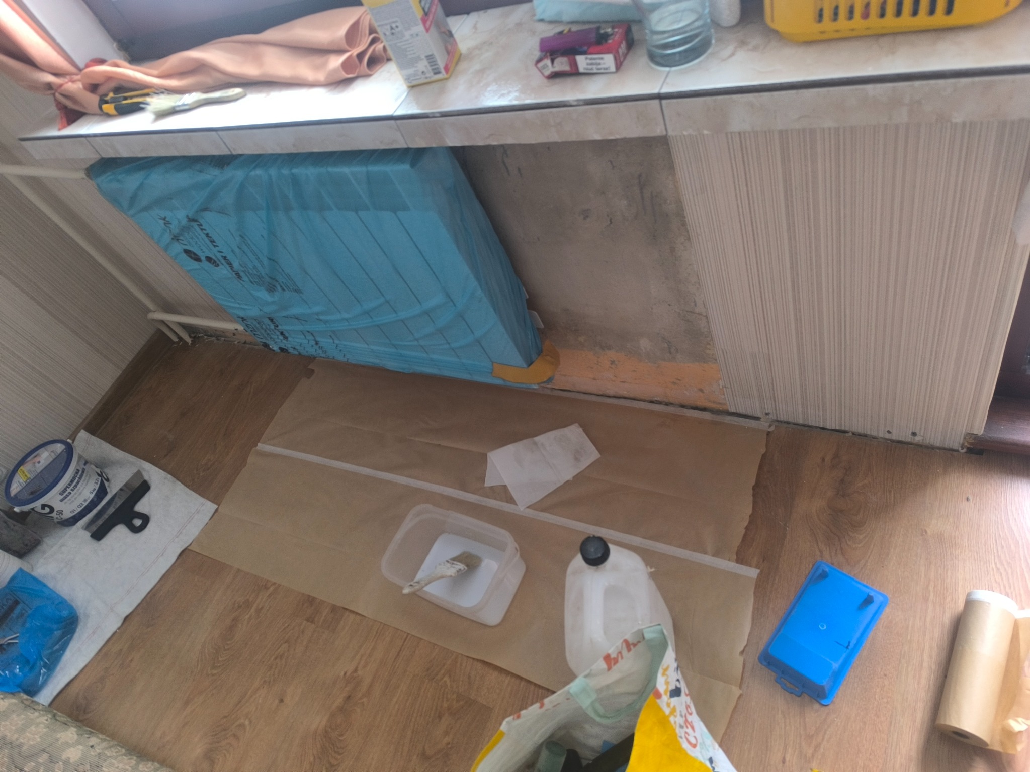 Przygotowania do malowania: zabezpieczone grzejniki, rozłożona folia ochronna, farba, pędzle i narzędzia na drewnianej podłodze. Widok z góry, wnętrze w trakcie remontu.