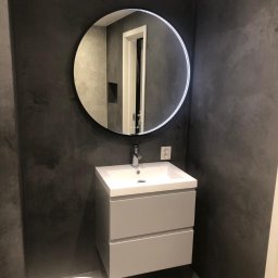 Marek Kemnitz - Wykończona łazienka z szarą ścianą, okrągłym lustrem z podświetleniem LED i białą umywalką z szafką. Minimalistyczny design, nowoczesne wykończenie.