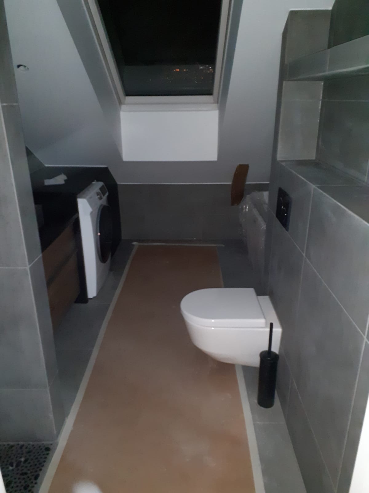 Nowoczesna łazienka na poddaszu z oknem dachowym. Szare płytki, biała toaleta podwieszana i pralka. Widoczny fragment podłogi zabezpieczony folią malarską.