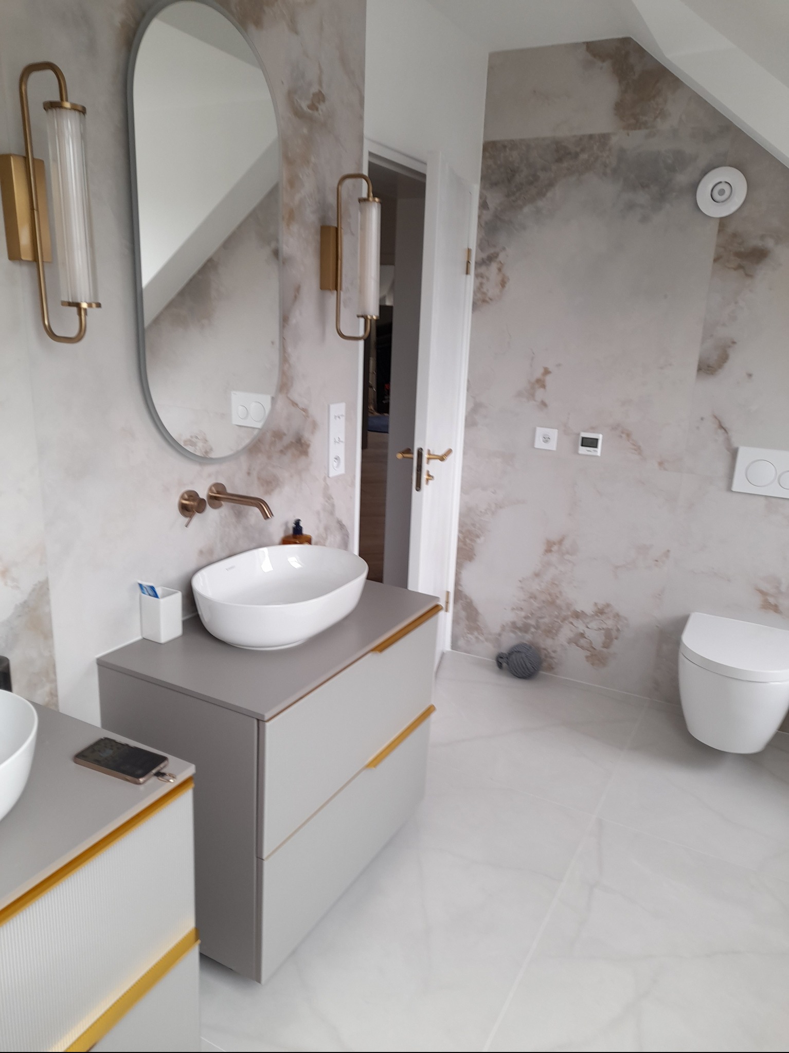 Jasna łazienka na poddaszu z marmurowymi ścianami, owalnym lustrem i umywalką nablatową na szarej szafce z złotymi akcentami. Widoczna toaleta i biała podłoga.