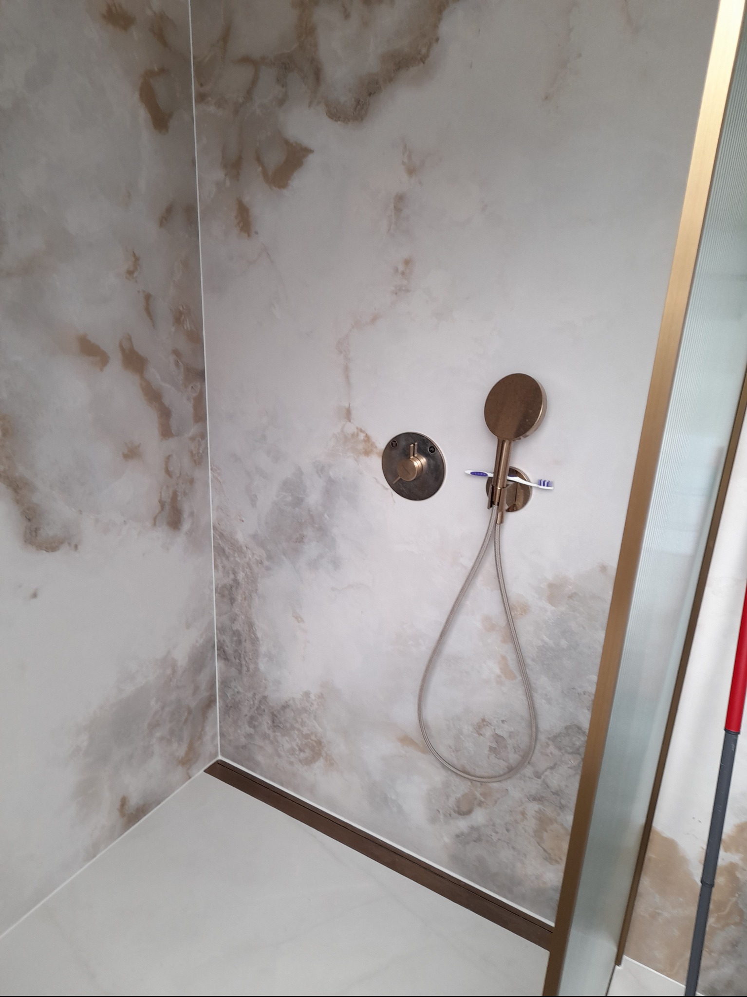 Wykończona kabina prysznicowa z marmurowymi płytami, złotą armaturą i szklanymi drzwiami. Widoczna szczoteczka do zębów na słuchawce prysznicowej.