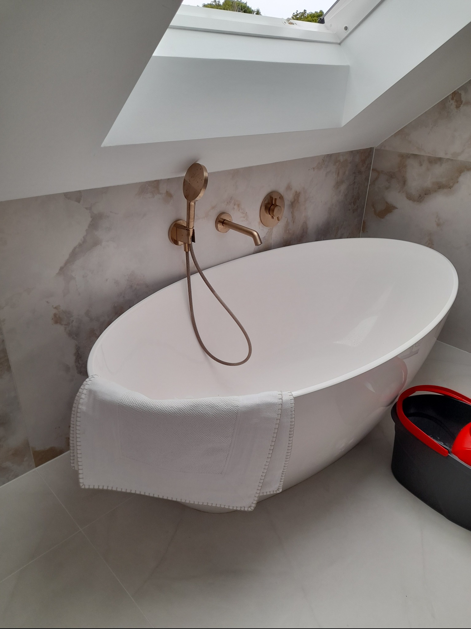 Nowoczesna, owalna wanna wolnostojąca z baterią w kolorze różowego złota, biały ręcznik i wiadro do mycia podłogi. Łazienka z oknem dachowym i marmurowymi płytkami.