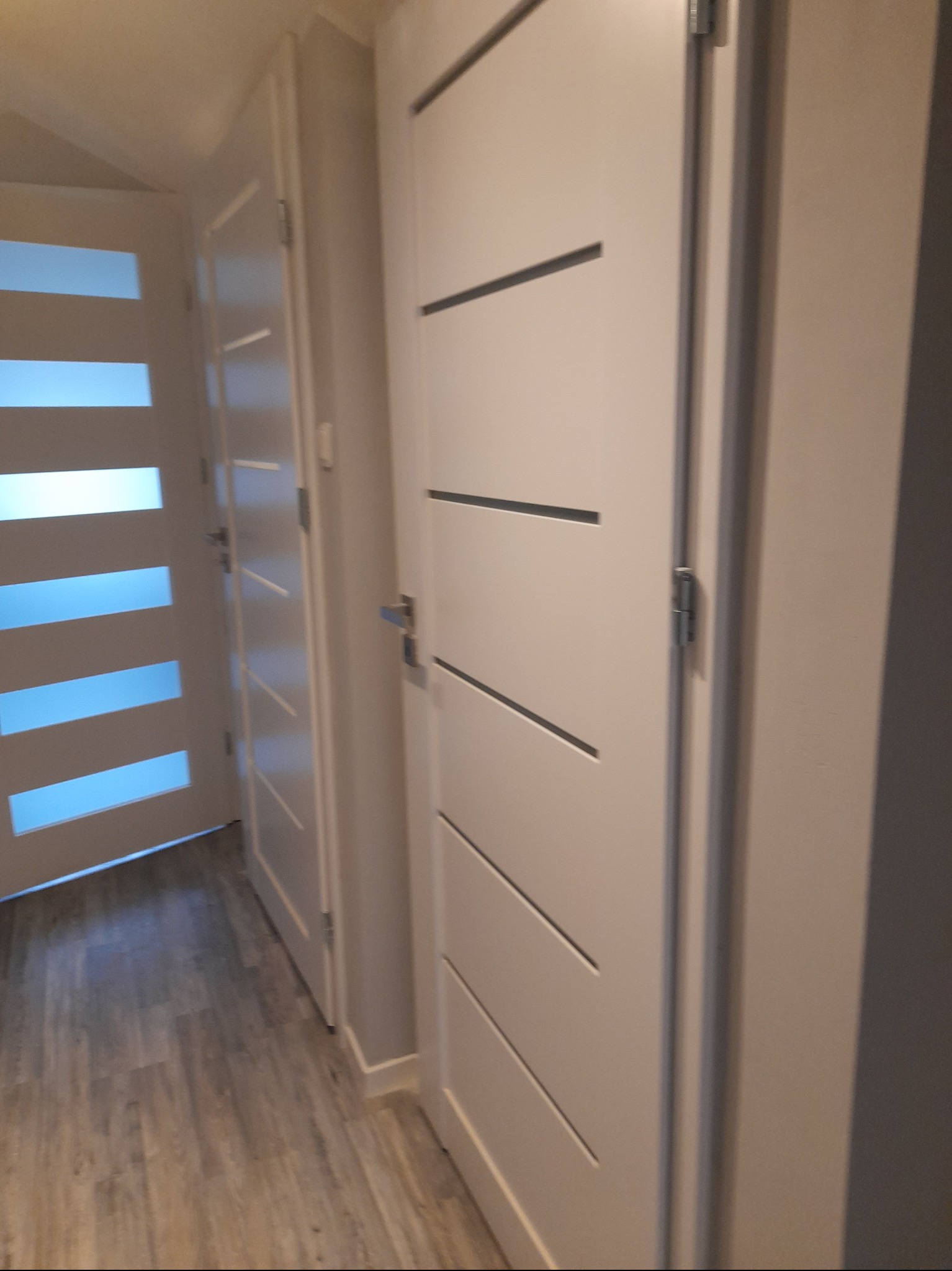 Białe, nowoczesne drzwi wewnętrzne z poziomymi frezami, zamontowane w jasnym pomieszczeniu. Widoczny fragment podłogi imitującej drewno i inne drzwi z przeszkleniami.