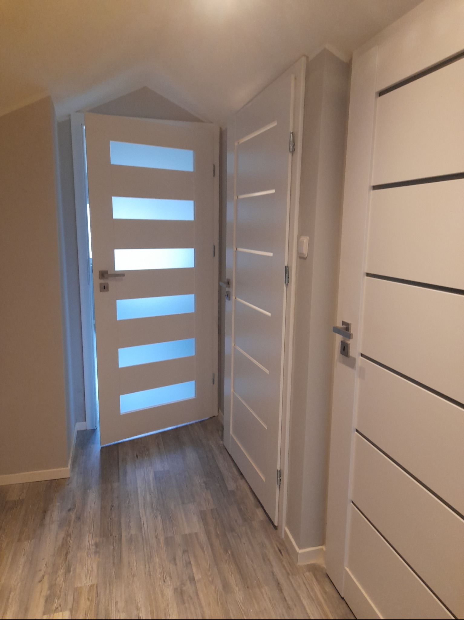 Trzy białe, nowoczesne drzwi wewnętrzne z poziomymi przeszkleniami i dekoracyjnymi frezami, zamontowane w jasnym korytarzu z panelami podłogowymi.