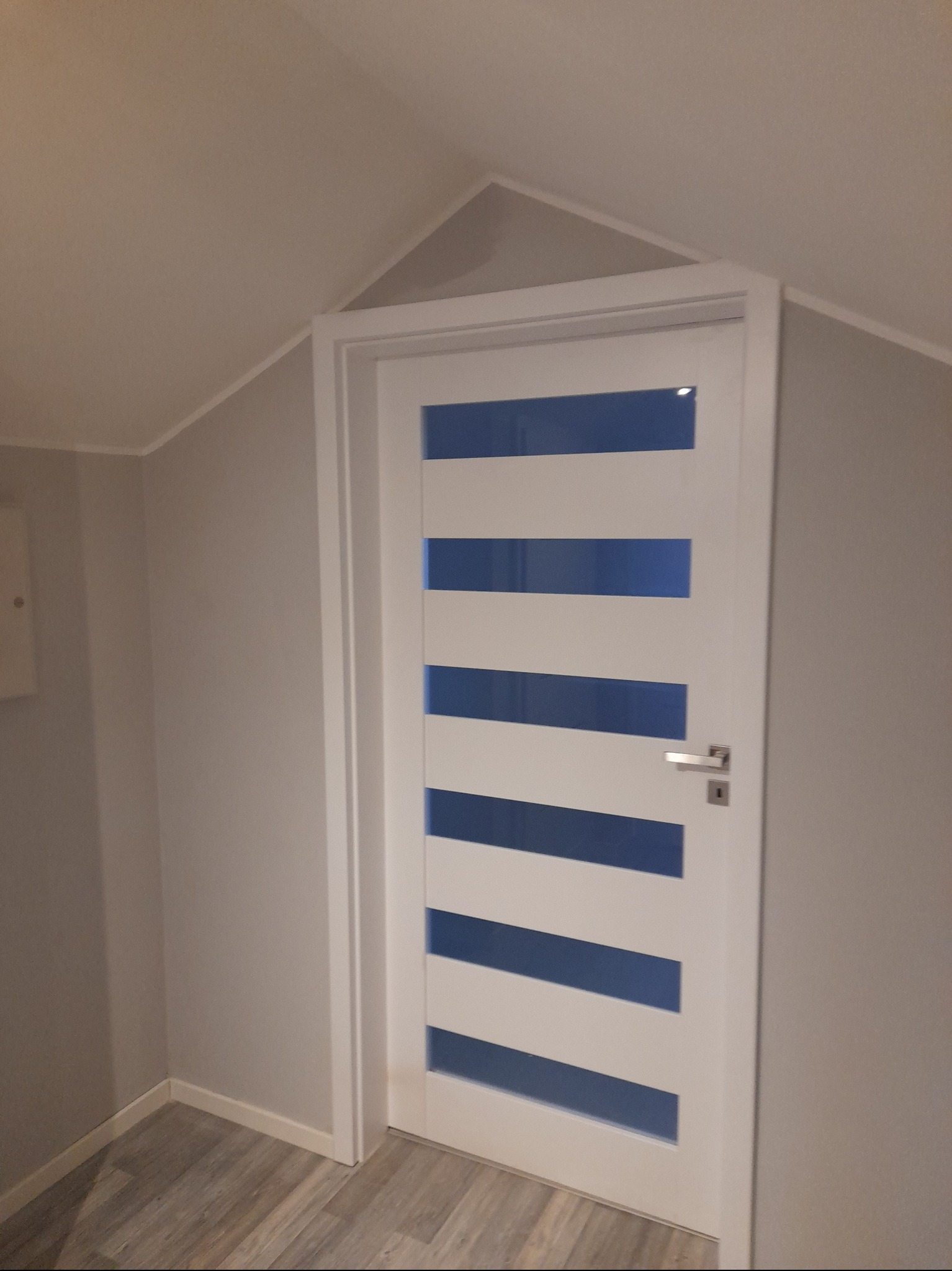 Białe drzwi z poziomymi, niebieskimi przeszkleniami, zamontowane w szarej ścianie na poddaszu. Widoczna klamka i fragment podłogi.