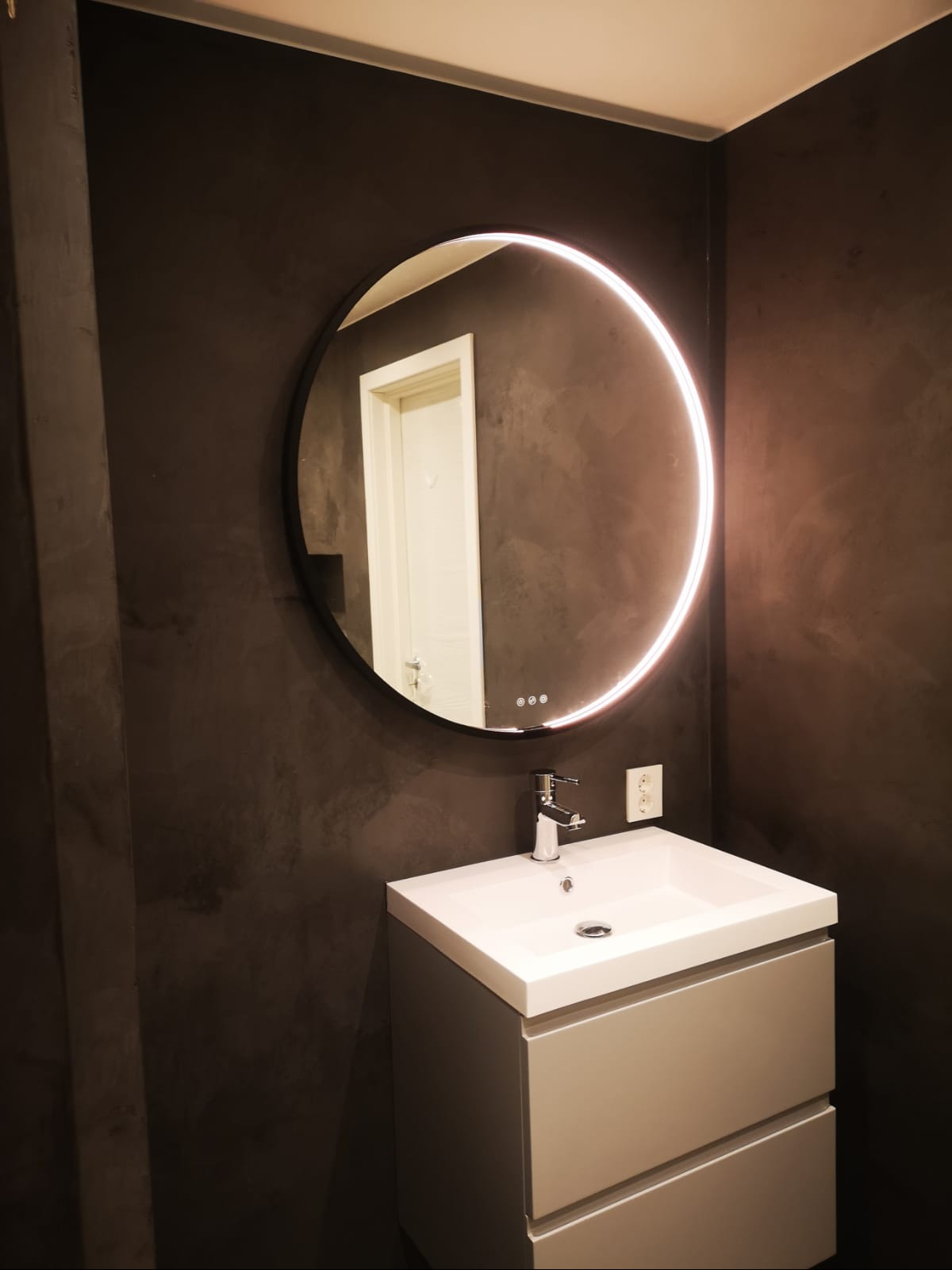 Nowoczesna łazienka z umywalką na szafce i okrągłym lustrem LED na ścianie w kolorze ciemnego grafitu z efektem dekoracyjnym. Widoczny fragment białych drzwi.