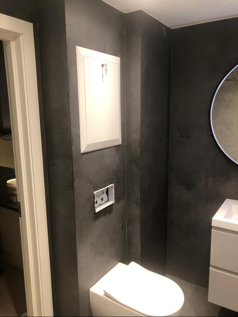 Nowoczesna łazienka z szarą ścianą o fakturze betonu, białą toaletą i umywalką. Widoczny otwór na gniazdko elektryczne i rewizja wodomierza.