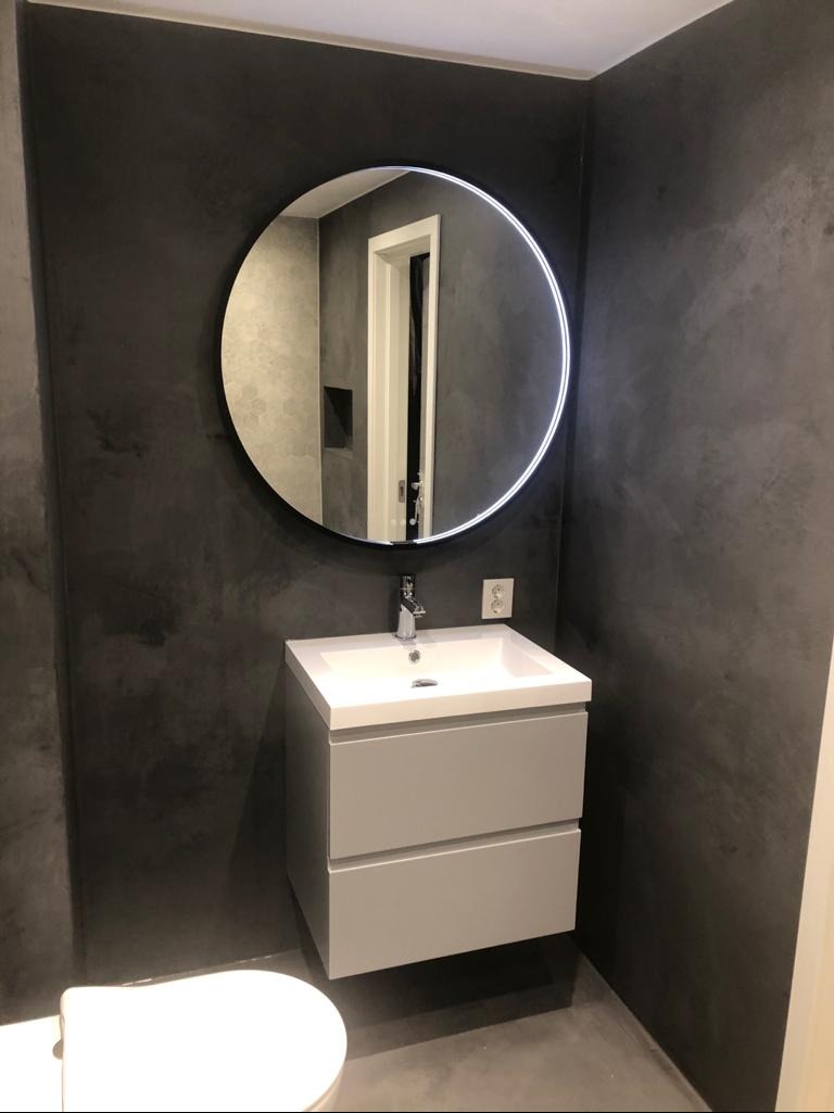 Wykończona łazienka z szarą ścianą, okrągłym lustrem z podświetleniem LED i białą umywalką z szafką. Minimalistyczny design, nowoczesne wykończenie.