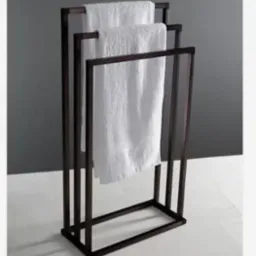 Stojak na ręczniki, metalowa konstrukcja w ciemnym kolorze z trzema białymi ręcznikami. Minimalistyczny design, widok z przodu na tle szaro-białej łazienki.