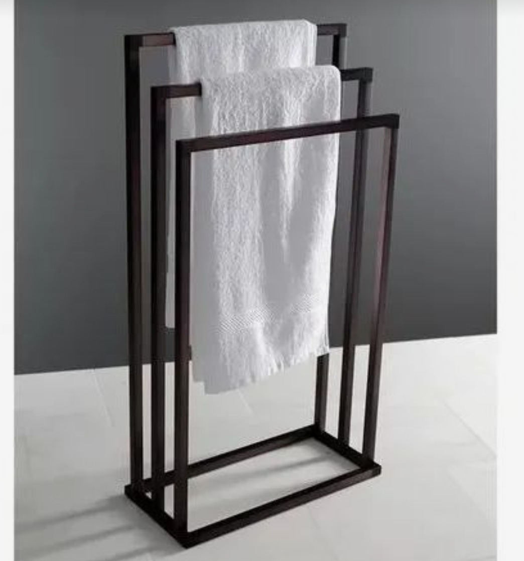 Stojak na ręczniki, metalowa konstrukcja w ciemnym kolorze z trzema białymi ręcznikami. Minimalistyczny design, widok z przodu na tle szaro-białej łazienki.