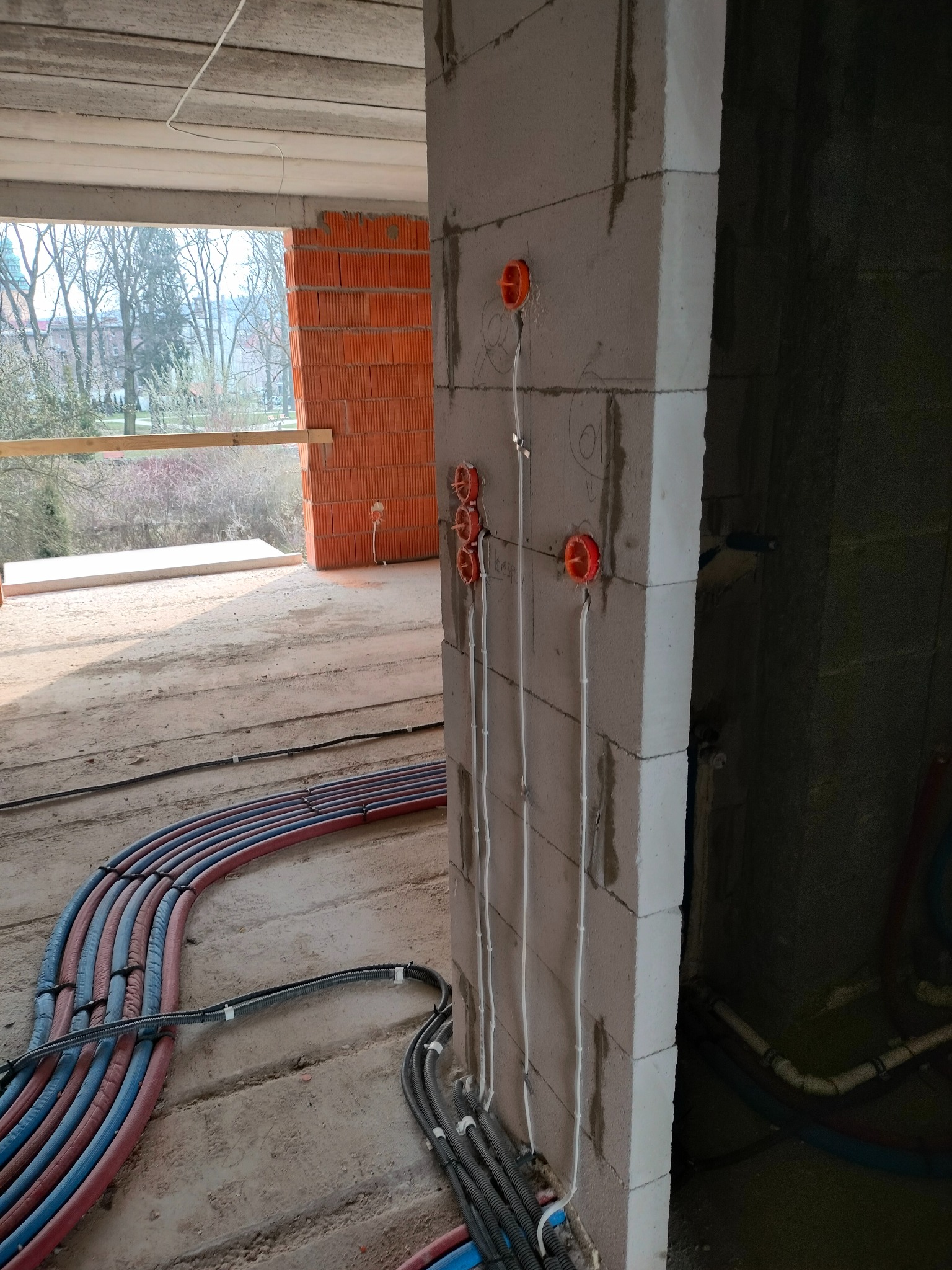 Wewnętrzne instalacje w surowym budynku: kable elektryczne wychodzące z pomarańczowych puszek na ścianie z betonu komórkowego, rury ogrzewania podłogowego na posadzce.