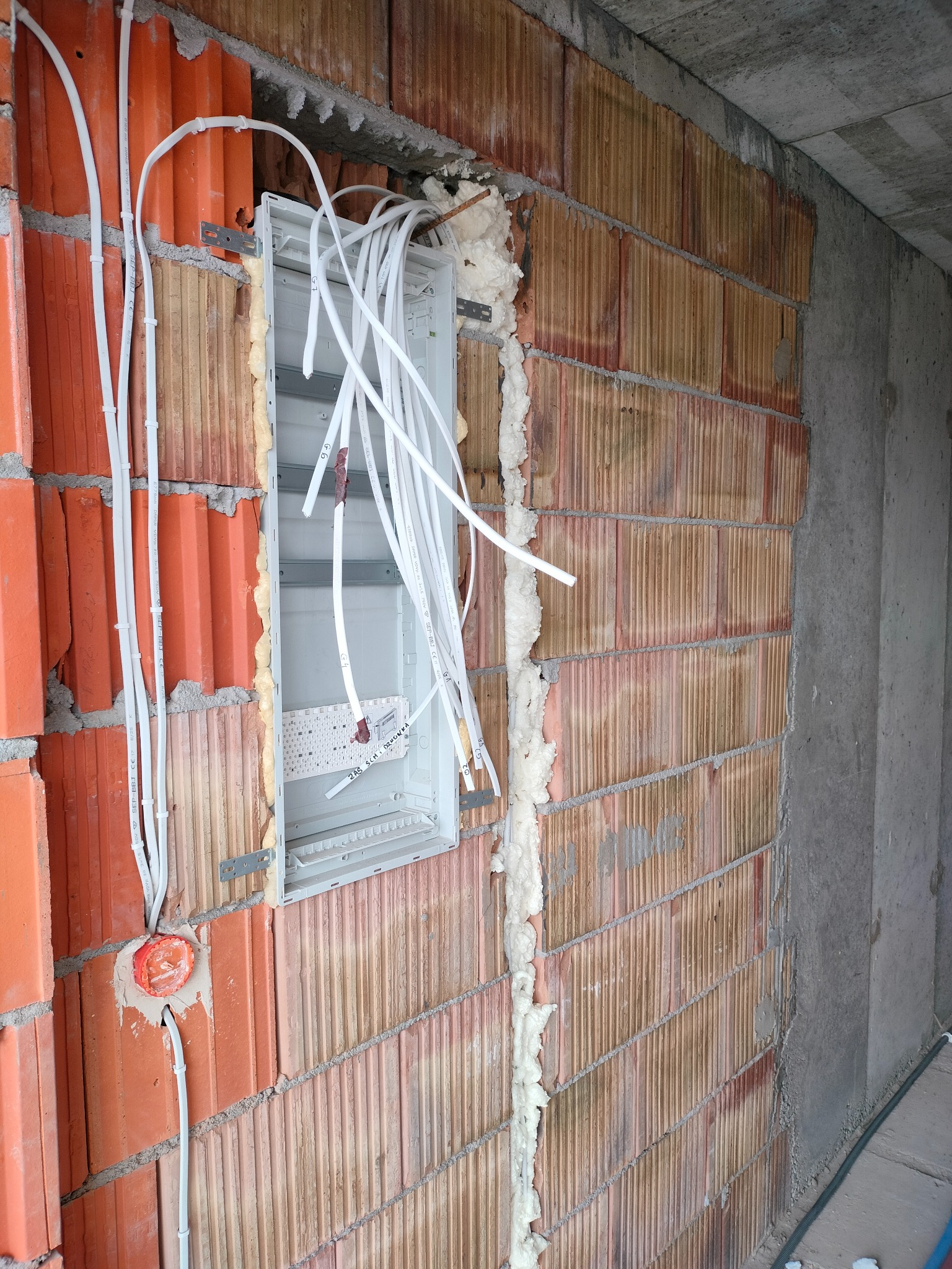 Instalacja elektryczna w trakcie montażu: puszka rozdzielcza z przewodami w ścianie z czerwonej cegły, zabezpieczona pianką montażową. Widoczne kable w peszlach.