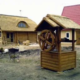 Studnia z drewna z kołowrotem i strzechą, w tle drewniana chata kryta strzechą i budynek z czerwonym dachem. Elementy dekoracyjne na posesji.