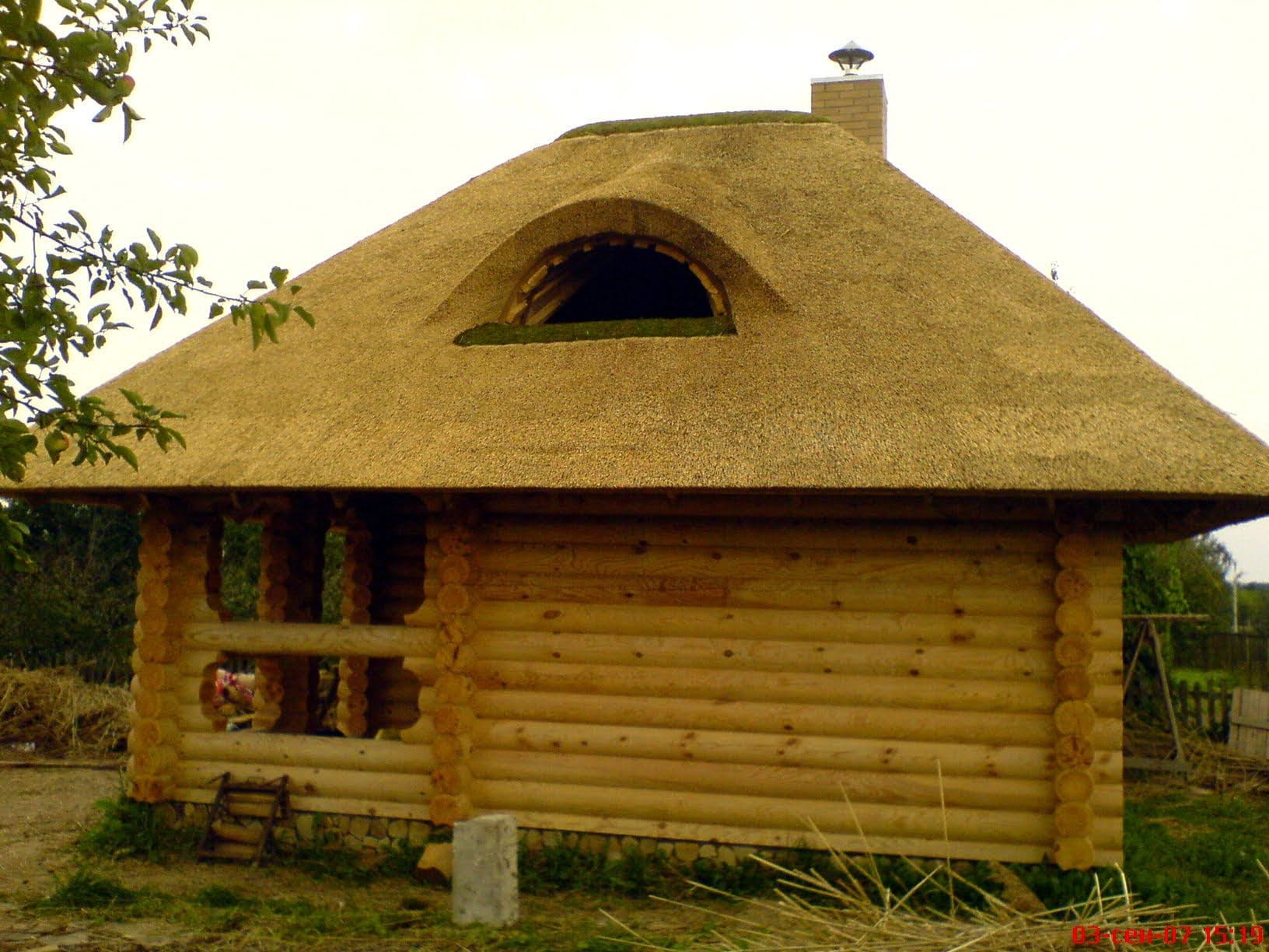 Drewniana altana z krytym strzechą dachem i oknem w kształcie oka, wykonana z bali. Widoczne detale konstrukcyjne i naturalne materiały.