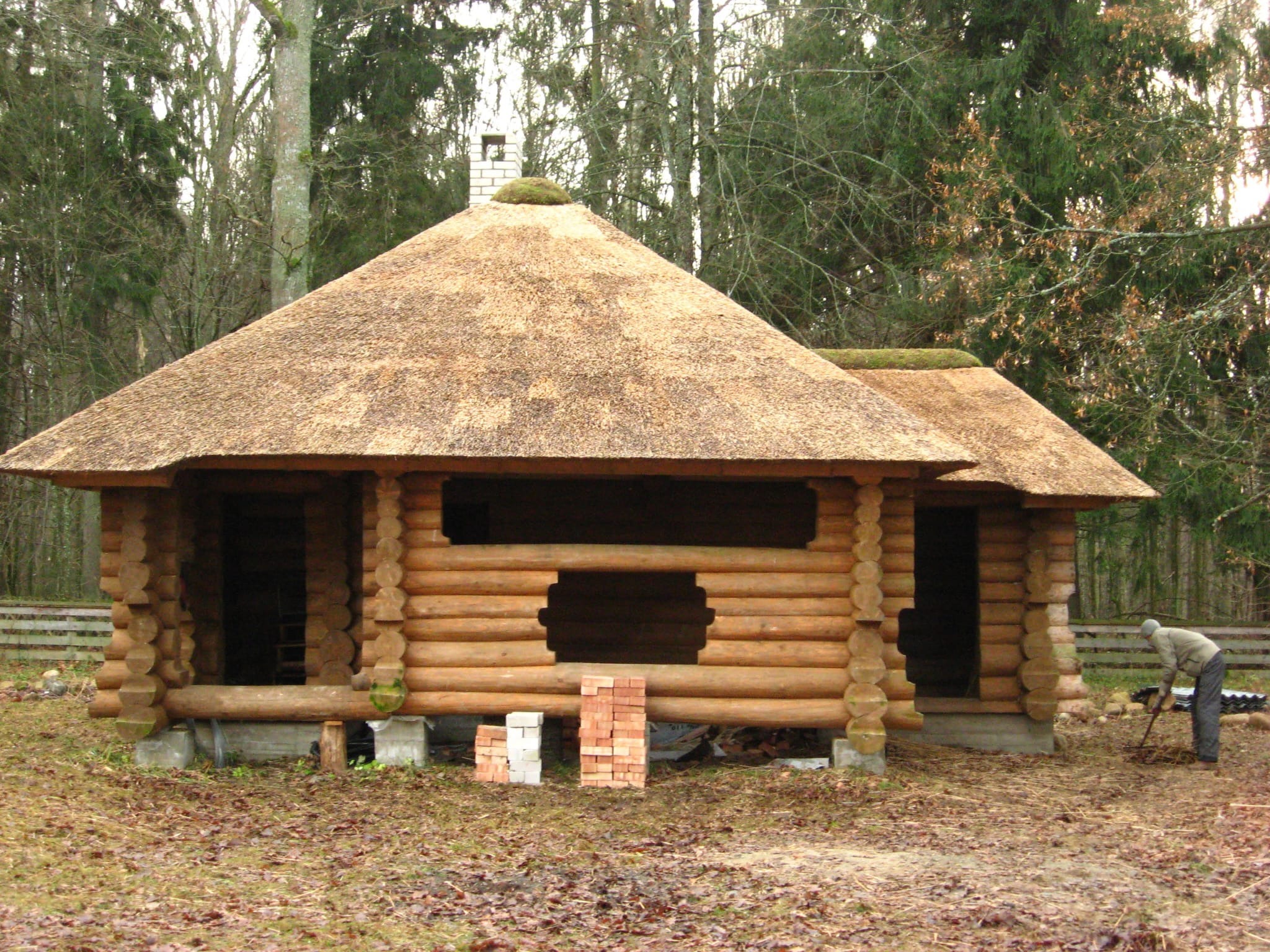 Wznoszona altana z bali drewnianych z nietypowym, strzechowym dachem. Obok sterta cegieł i pracownik porządkujący teren. W tle las.