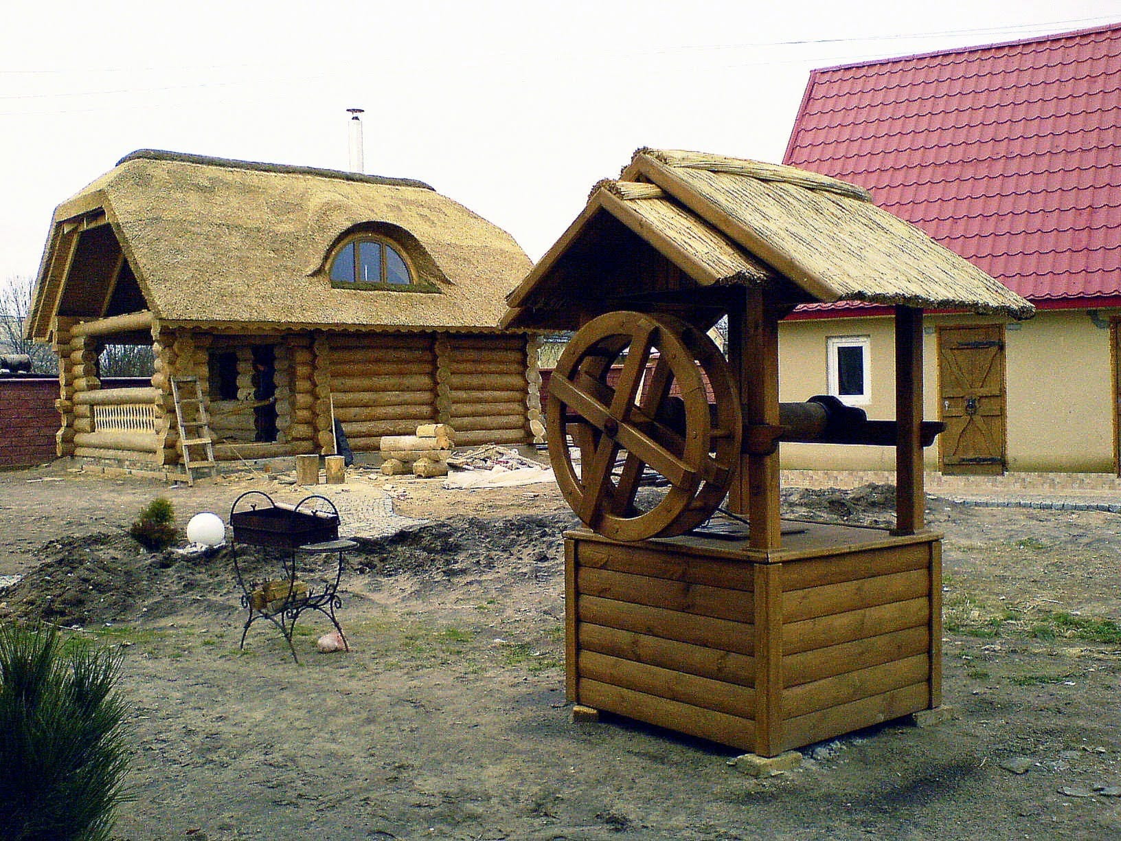 Studnia z drewna z kołowrotem i strzechą, w tle drewniana chata kryta strzechą i budynek z czerwonym dachem. Elementy dekoracyjne na posesji.