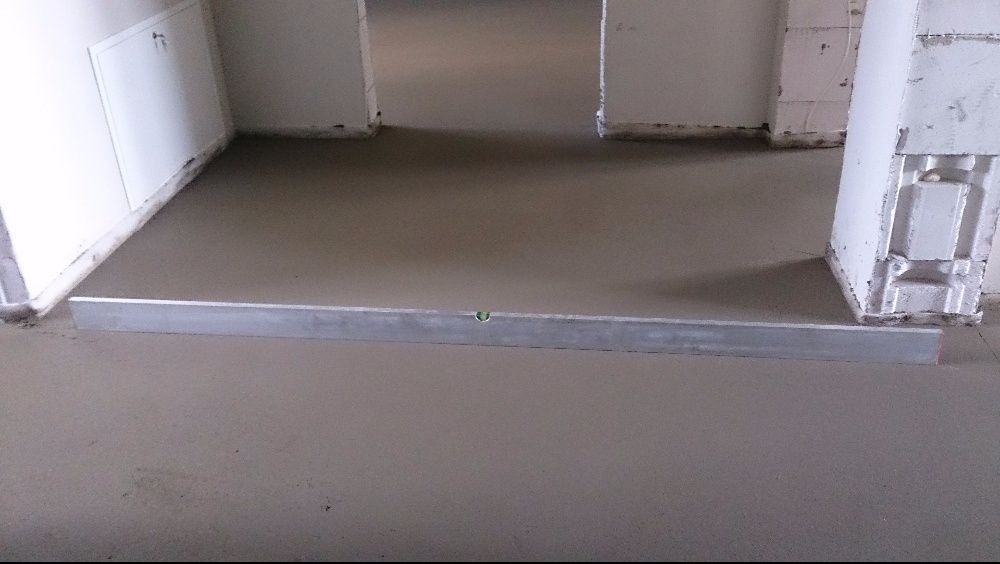 Wyrównywanie świeżo wylanej posadzki poziomnicą w Michałowie. Widoczne ściany w surowym stanie i fragment wylewki w tle.