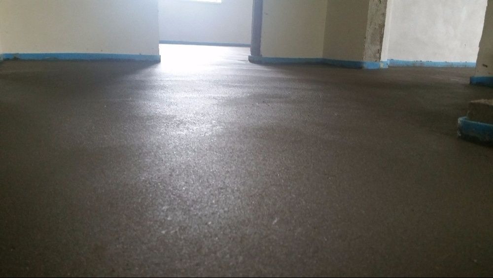 Świeżo wylana, gładka posadzka z jastrychu, zabezpieczona niebieską taśmą przy ścianach w jasnym, surowym wnętrzu. Widoczne refleksy światła.