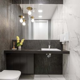 Maximus - Nowoczesna łazienka z białymi marmurowymi płytkami, czarną szafką pod umywalką i złotymi lampami wiszącymi. Widoczne lustro i toaleta.