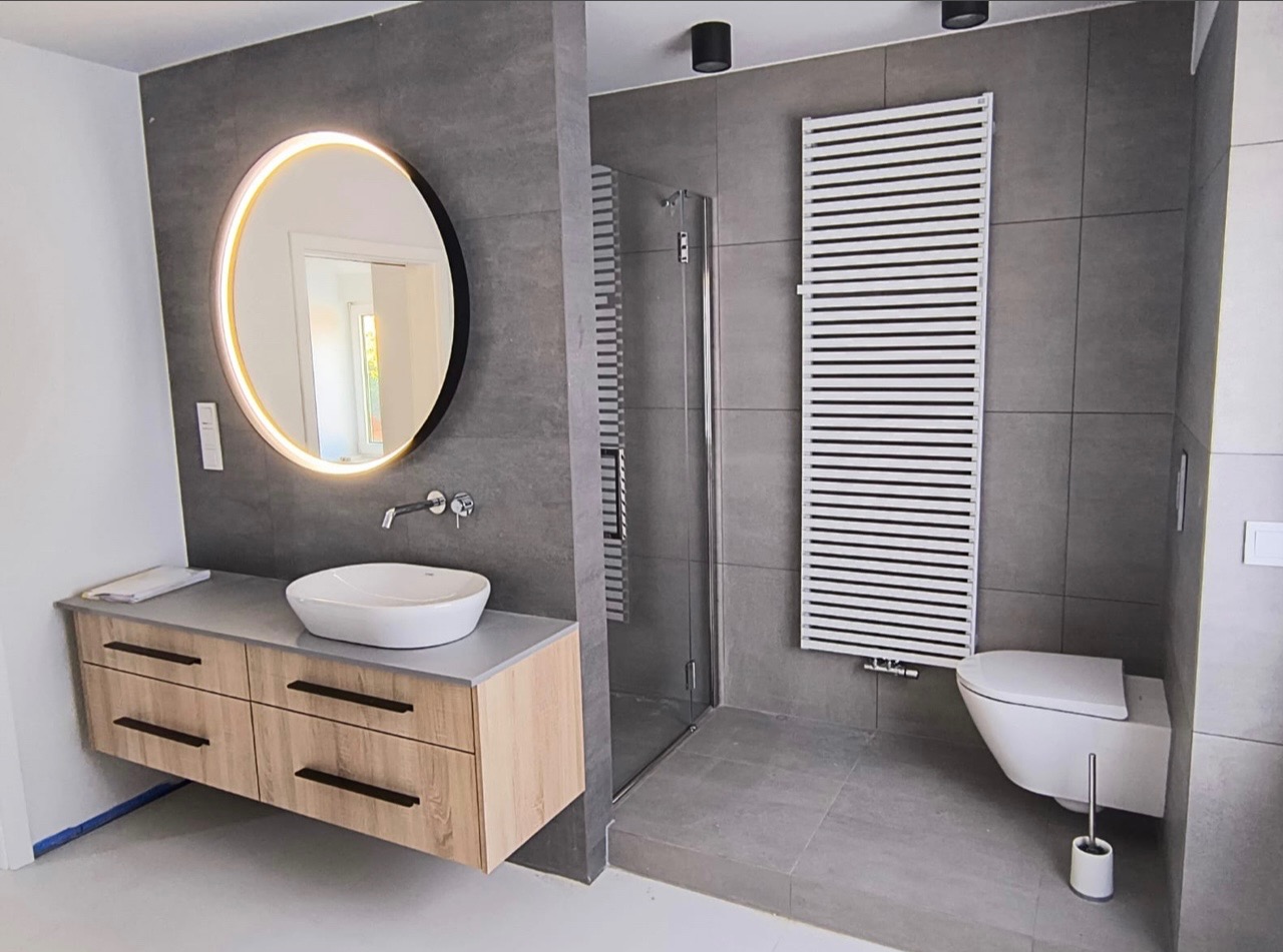 Nowoczesna łazienka z szarymi płytkami, okrągłym lustrem z podświetleniem LED, drewnianą szafką pod umywalką i grzejnikiem drabinkowym.