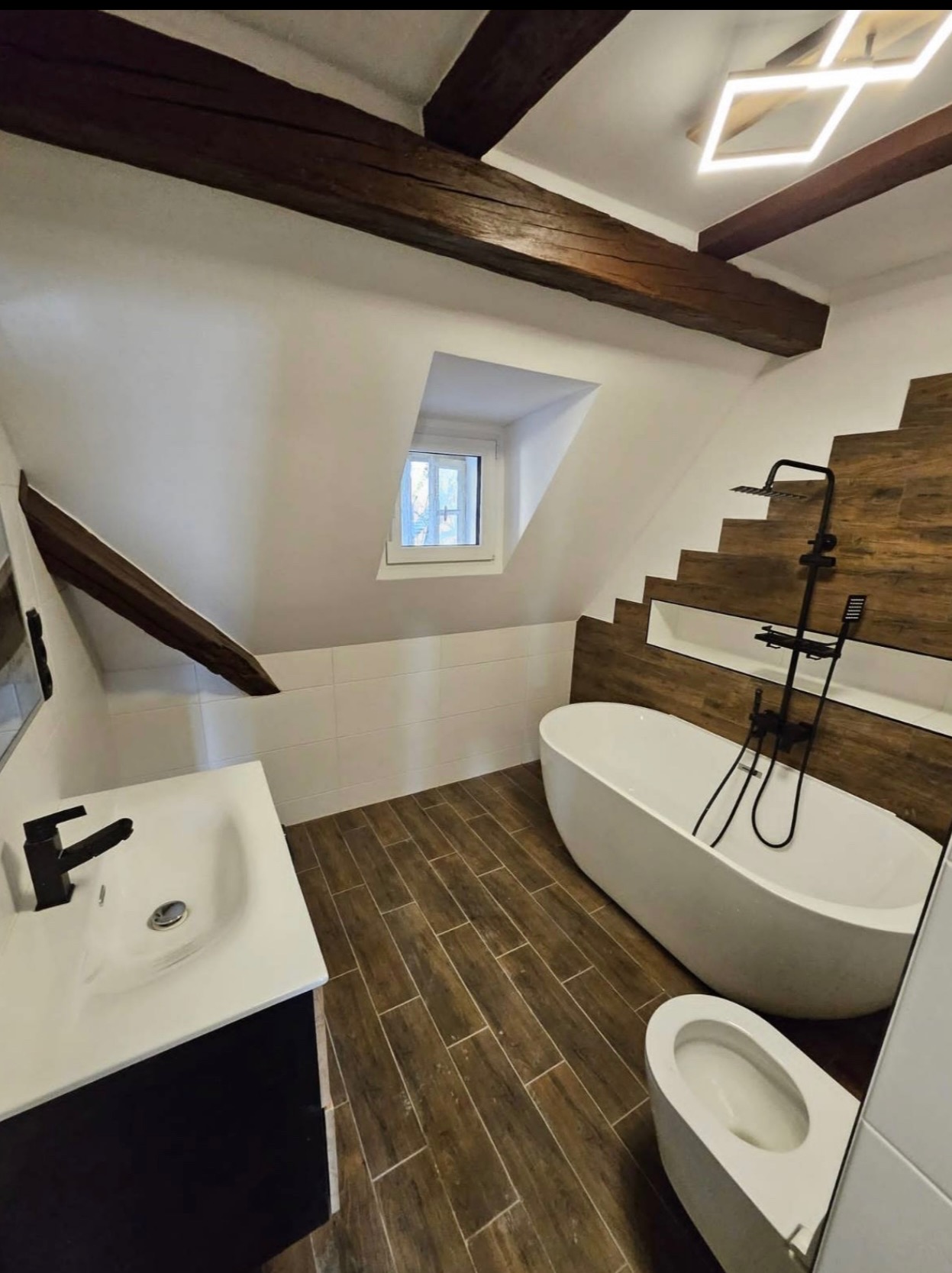 Stylowa łazienka na poddaszu z drewnianymi belkami, białą wanną wolnostojącą i armaturą w kolorze czarnym. Uwagę przykuwa oryginalna ściana z drewnopodobnych płytek.