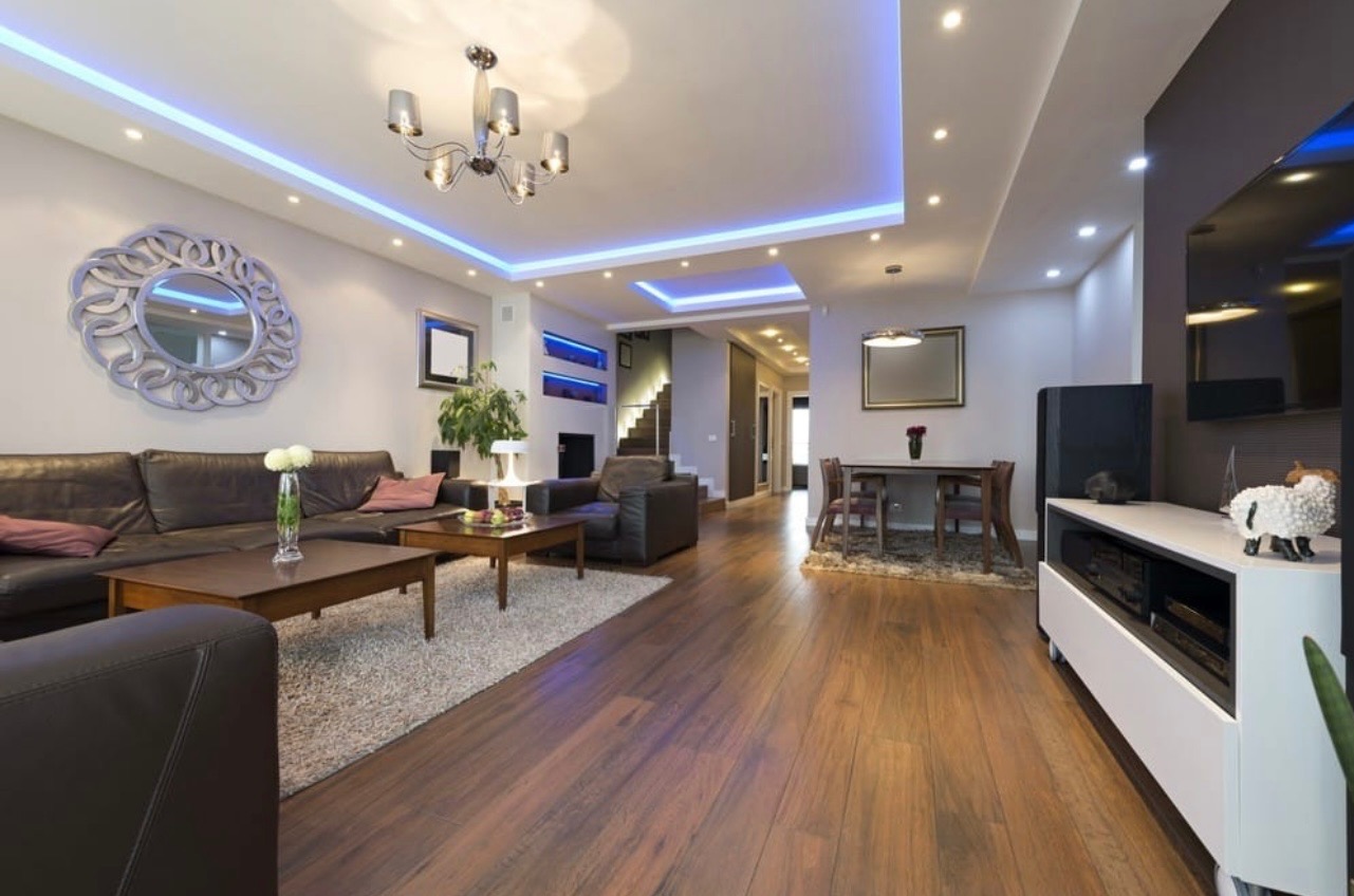 Elegancki salon z drewnianą podłogą, skórzaną sofą i dekoracyjnym lustrem. Podwieszany sufit z niebieskim oświetleniem LED tworzy nowoczesny klimat wnętrza.
