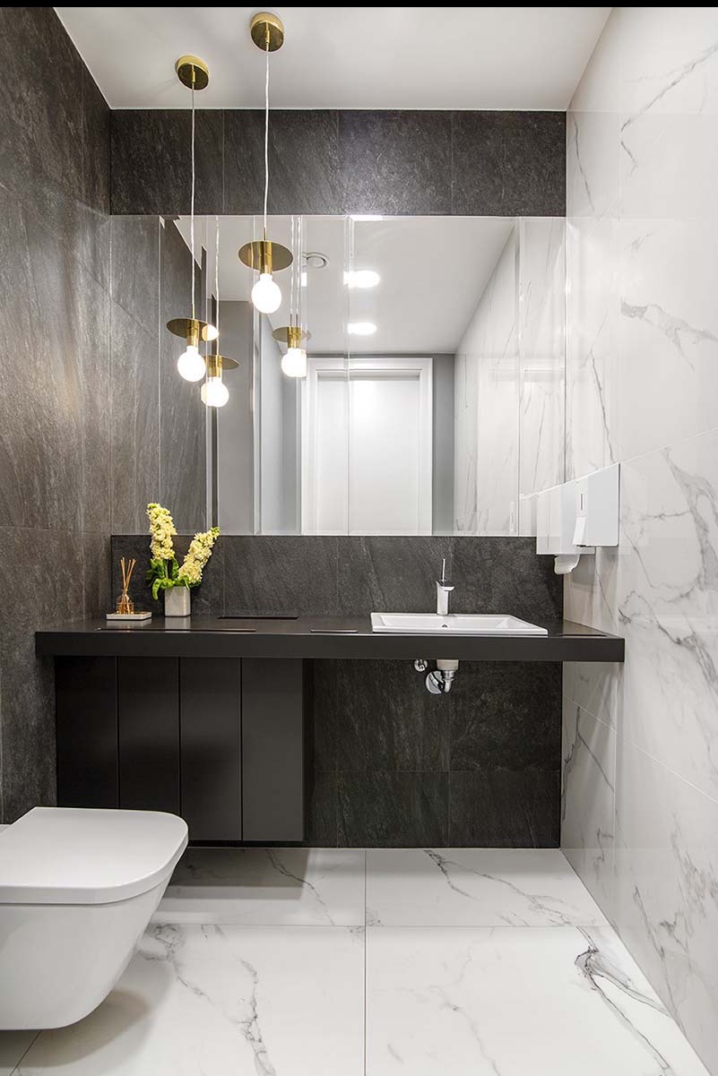 Nowoczesna łazienka z białymi marmurowymi płytkami, czarną szafką pod umywalką i złotymi lampami wiszącymi. Widoczne lustro i toaleta.