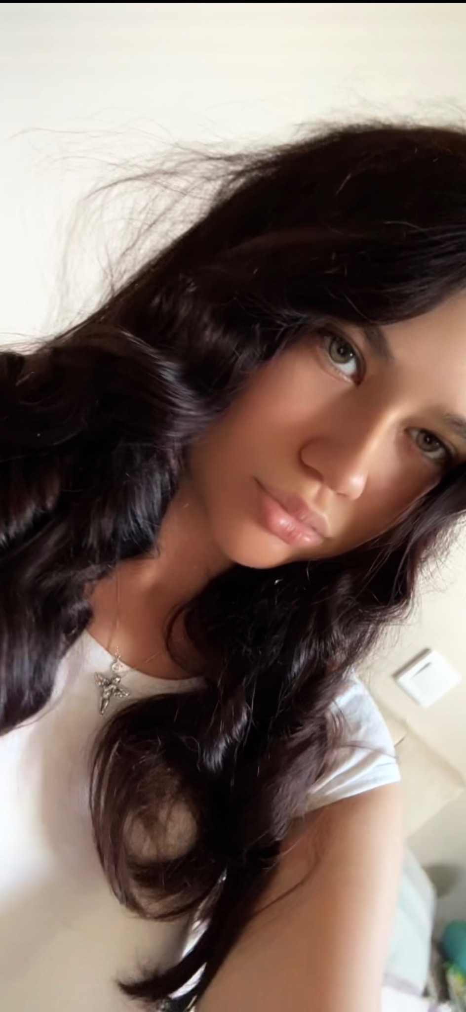 Portret uśmiechniętej niani z Warszawy, z długimi, kręconymi włosami i delikatnym makijażem, w białej koszulce i z srebrnym naszyjnikiem.