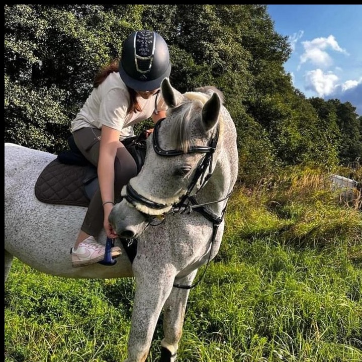 Dziewczyna w kasku jeździeckim siedzi na siwym koniu na tle zieleni. Koń ma na sobie uzdę z futerkiem. Uwagę przykuwa specyficzne umaszczenie konia.