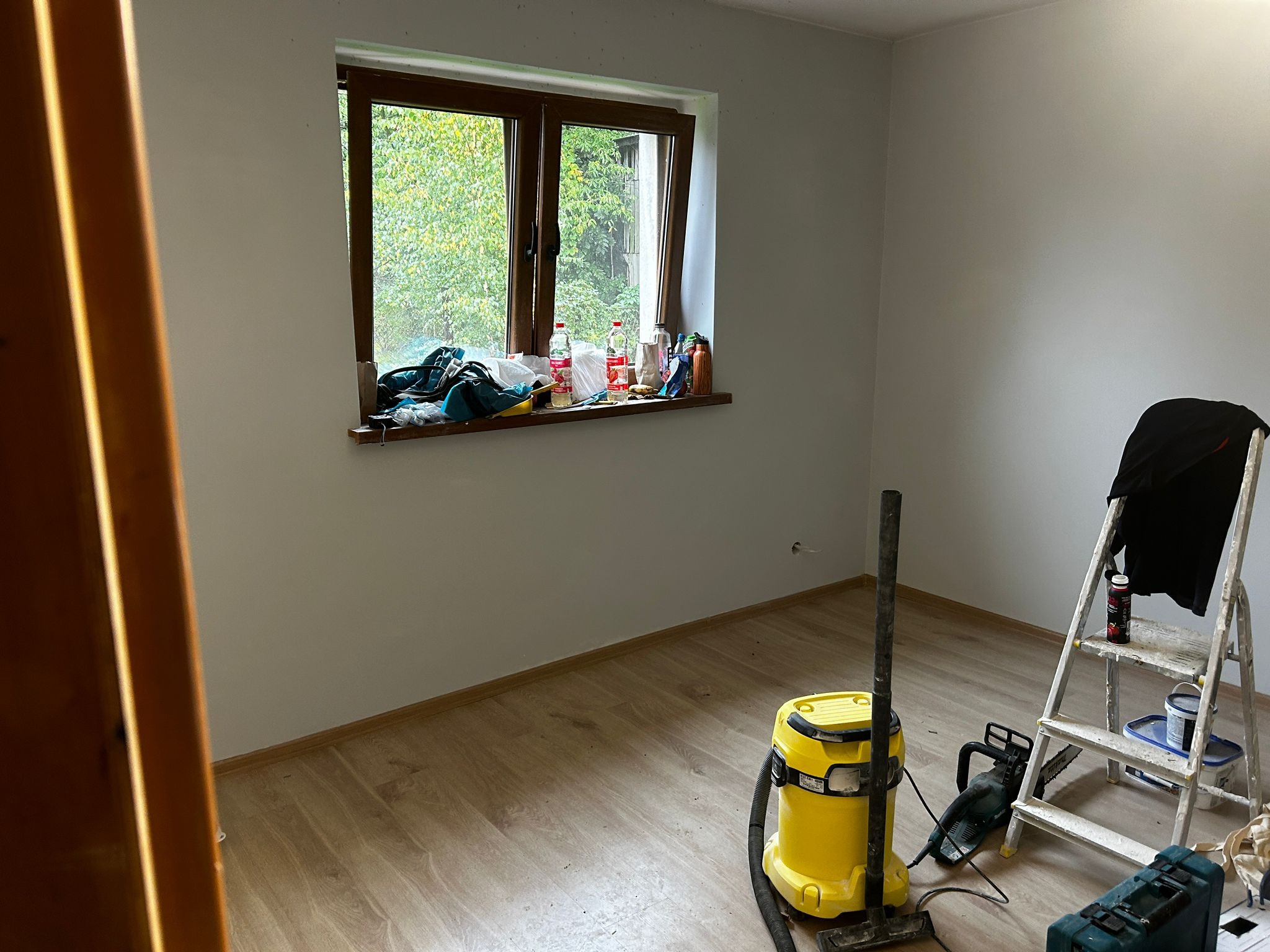 Pokój po remoncie: świeżo pomalowane ściany, okno z widokiem na zieleń, drabina, odkurzacz i narzędzia na podłodze. Widać, że prace malarskie zostały zakończone.