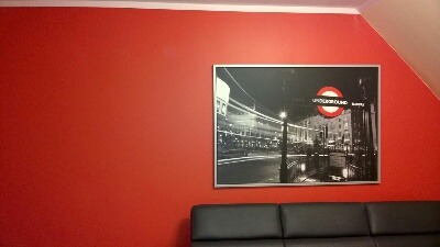 Czerwona ściana w pokoju na poddaszu, ozdobiona czarno-białym plakatem z motywem londyńskiego metra. Widoczny fragment czarnej sofy. Oświetlenie dzienne.