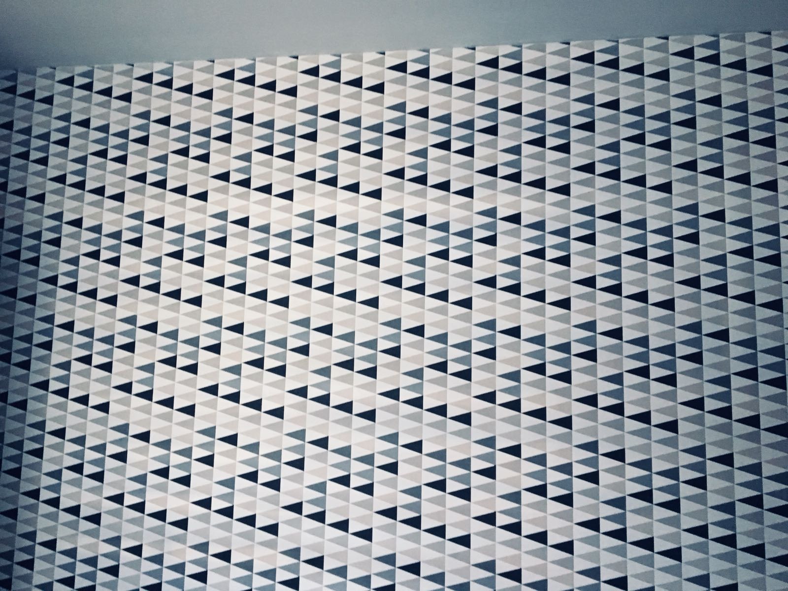 Ściana pokryta wzorzystą tapetą w trójkąty w odcieniach szarości i bieli, tworząca nowoczesny, geometryczny design wnętrza. Widoczny fragment sufitu.