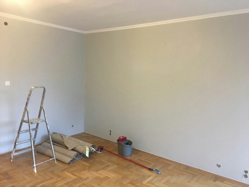 Pokój po malowaniu na szaro z listwą sufitową. Na podłodze parkiet, stoi drabina, wiadro z farbą i wałki malarskie. Widoczne gniazdka elektryczne.