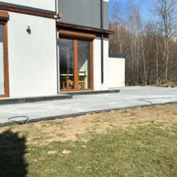 Nowoczesny taras z szarej kostki brukowej przy domu z drewnianą stolarką okienną. Widoczny wykop i łopata, sugerujące prace wykończeniowe.