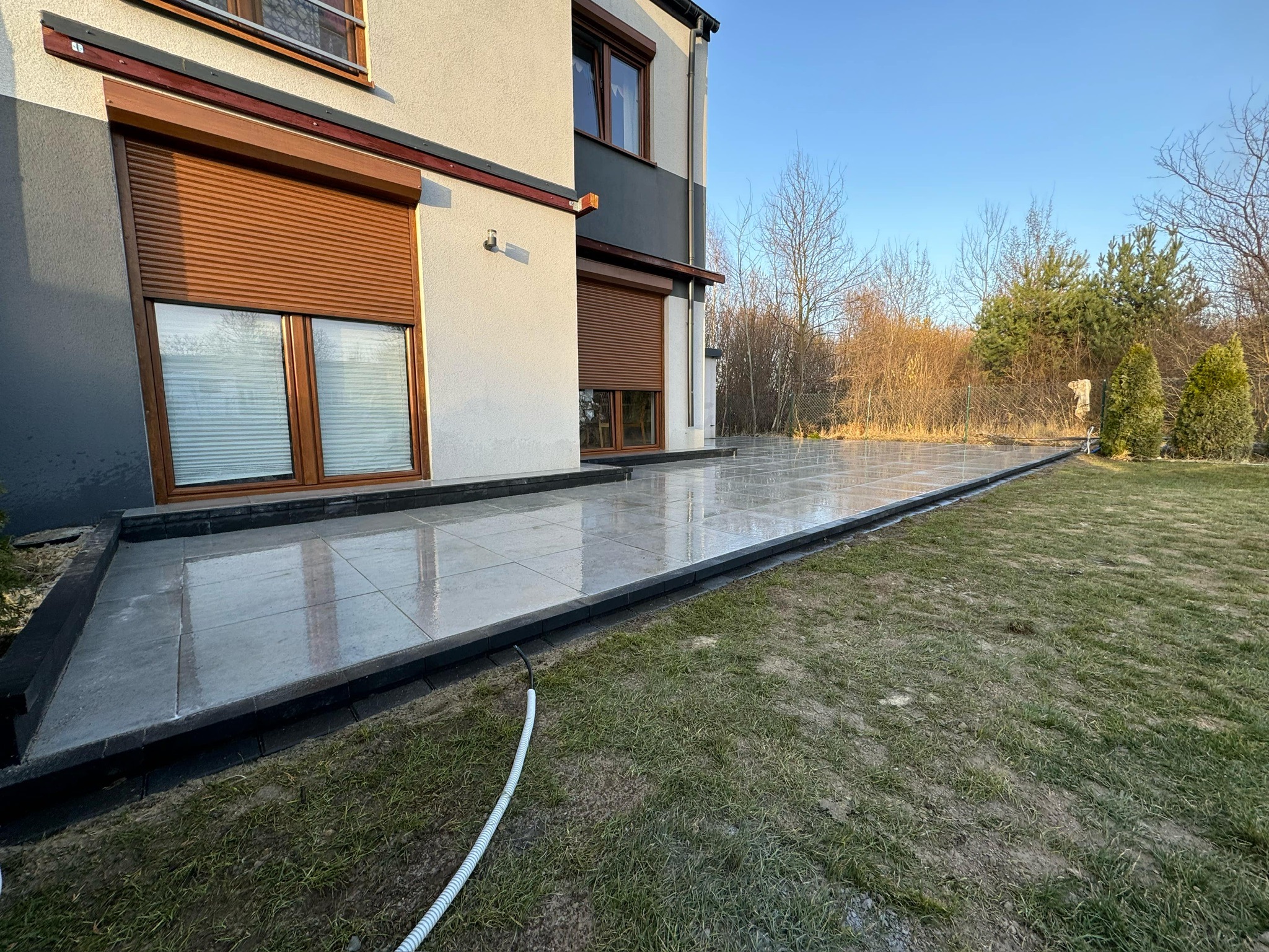 Jasnoszara kostka brukowa na tarasie przylegającym do domu z brązowymi roletami. Widoczne trawnik i drzewa w tle, biały wąż ogrodowy leży na trawie.