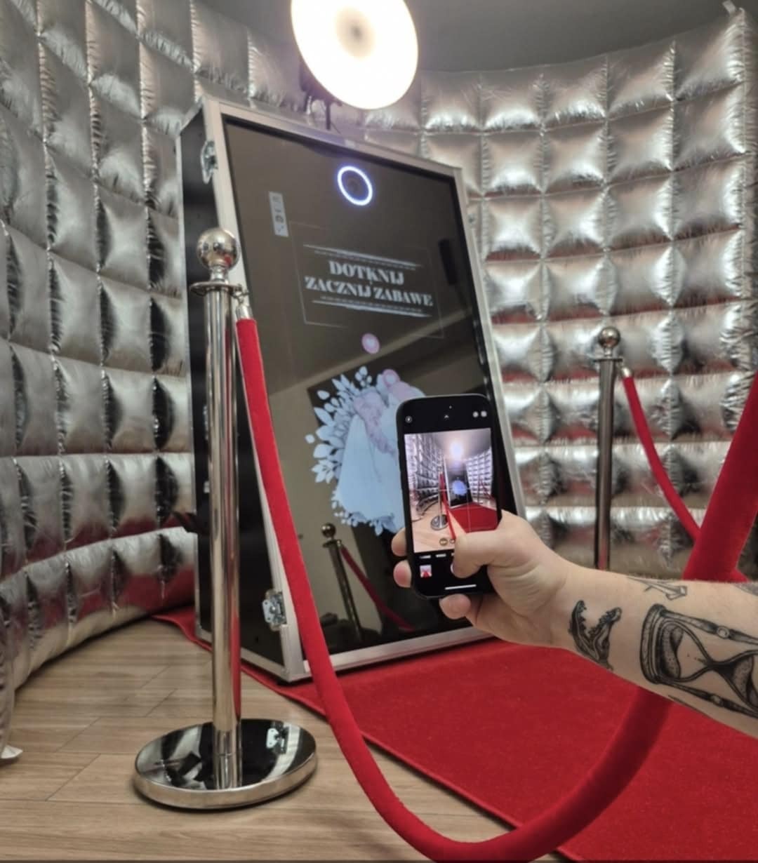 Osoba fotografuje telefonem fotobudkę z lustrem, otoczoną srebrną ścianką i czerwonym dywanem. Na ekranie fotobudki napis: Dotknij i zacznij zabawę.