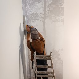 Ruslan Kostenko - Pracownik na drabinie w Poznaniu wygładza szpachlą ścianę z motywem leśnym. Widoczne narzędzia i fragment taśmy malarskiej. Praca w toku, wnętrze.