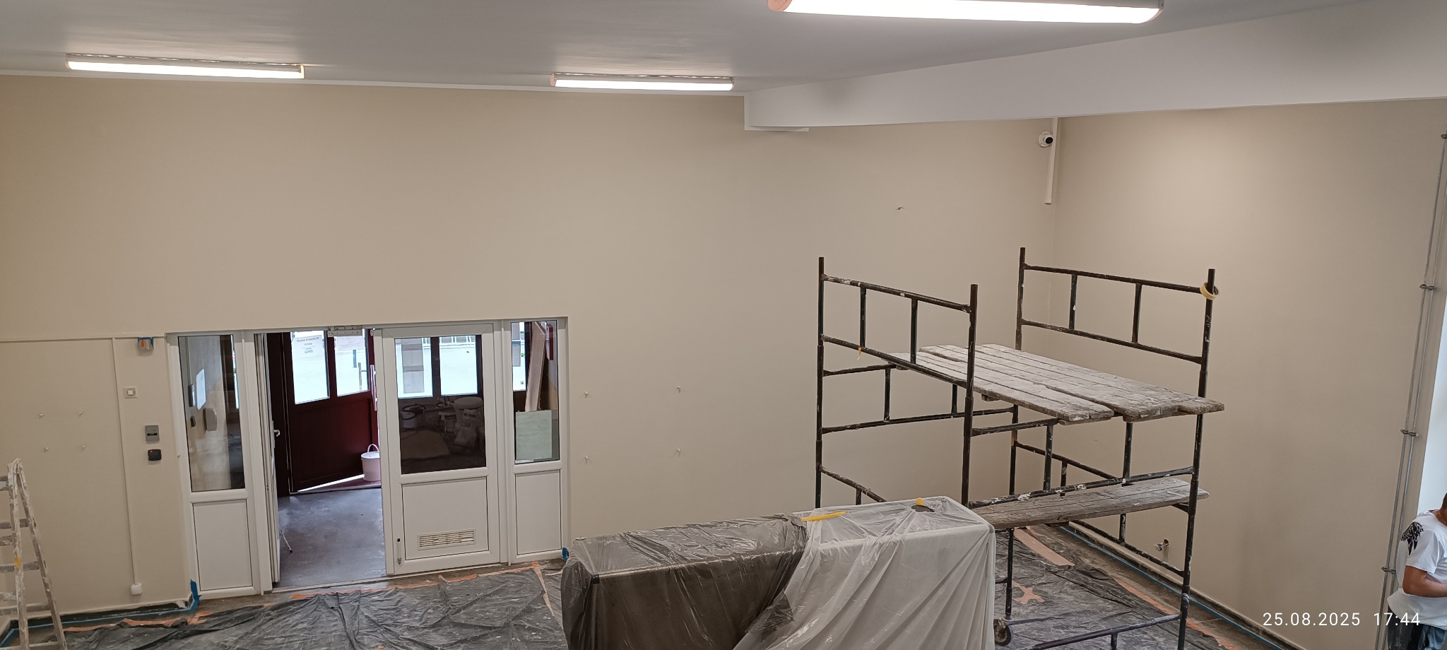 Wnętrze po malowaniu: kremowe ściany, rozstawione rusztowania, zabezpieczone folią meble. Widoczne ślady po szpachlowaniu. Oświetlenie sufitowe.