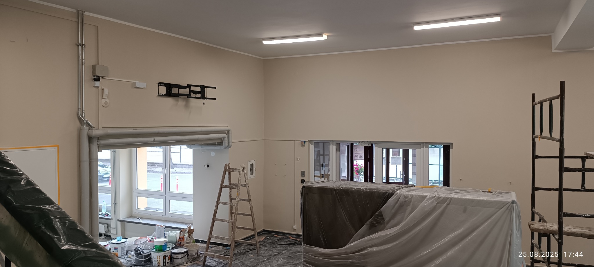 Wnętrze po malowaniu ścian na beżowo. Widoczne zakryte meble, drabina, puszki z farbą i rusztowanie. Ściany gładkie, z zamontowanym uchwytem na telewizor.