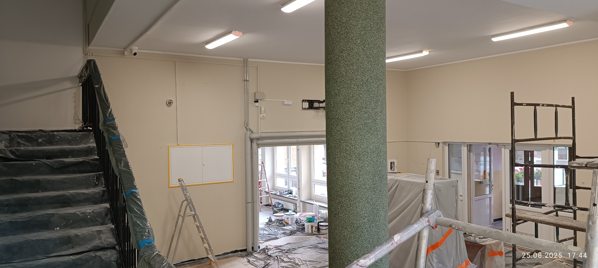 Klatka schodowa w trakcie malowania: zabezpieczone schody folią, drabina oparta o ścianę, widoczne puszki z farbą. Ściany w jasnym odcieniu beżu, kolumna w zielonej okleinie.