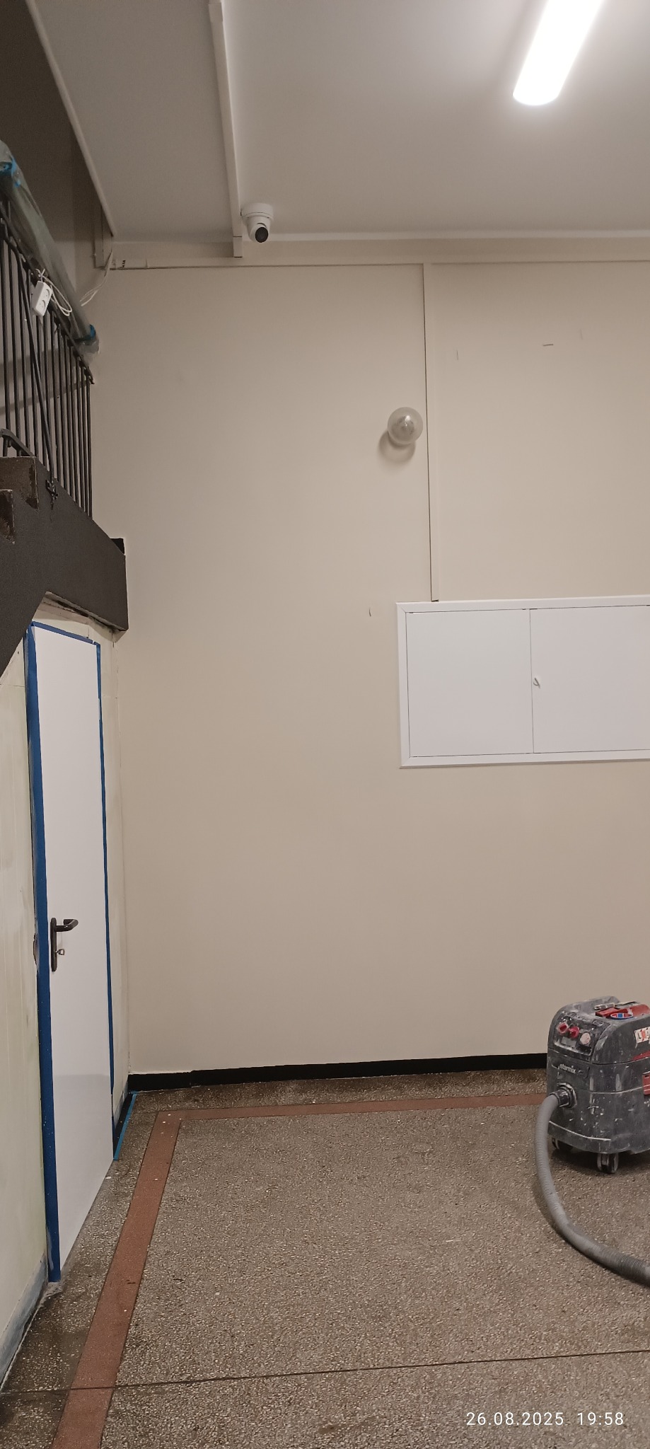 Wnętrze po malowaniu: jasne ściany, białe drzwi z niebieskim zabezpieczeniem, widoczny sprzęt do szlifowania. Minimalistyczne oświetlenie i czujnik na suficie.