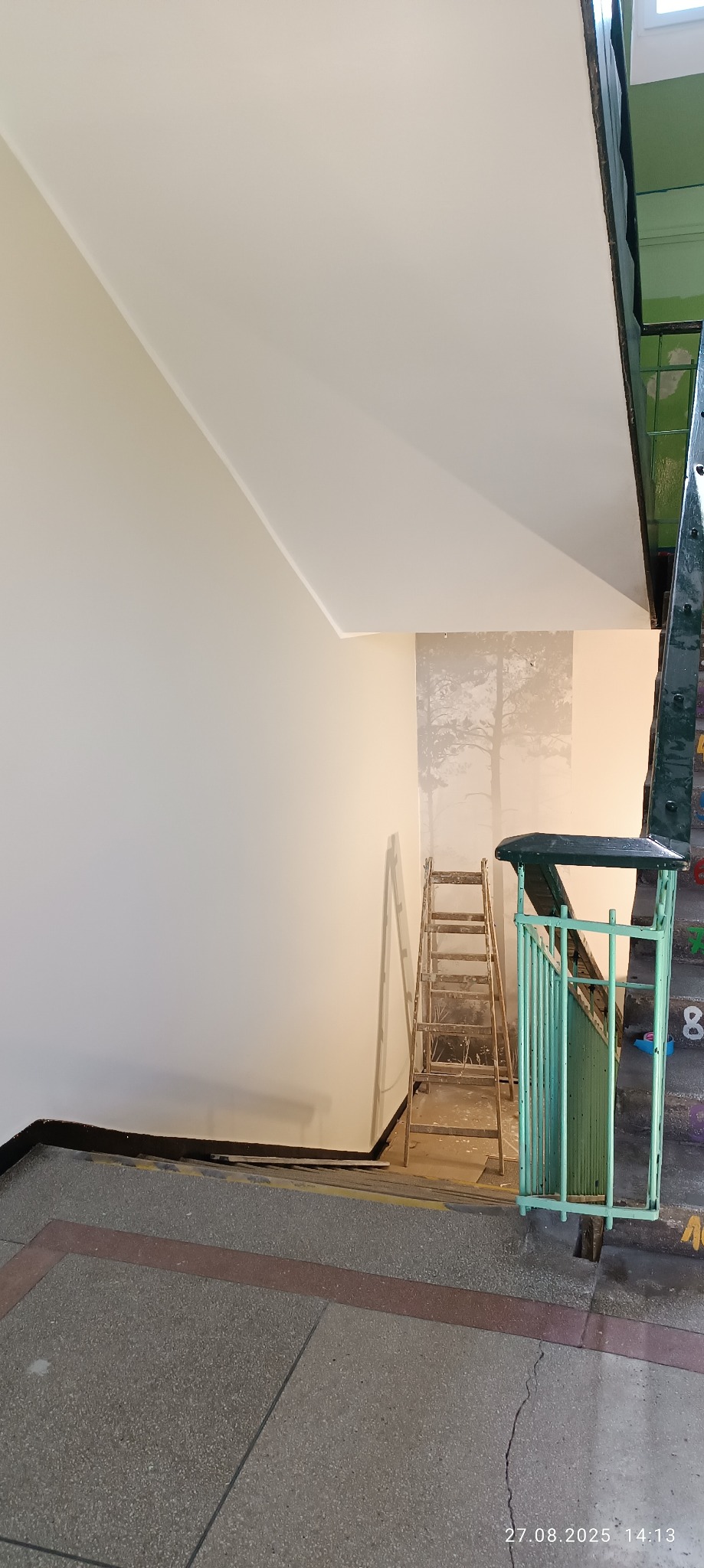 Klatka schodowa w trakcie malowania: świeżo pomalowana ściana kontrastuje z fragmentem przed malowaniem. Drewniana drabina oparta o ścianę. Widoczne schody i fragment balustrady.