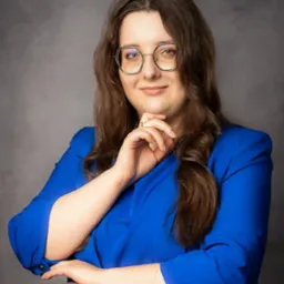 Portret kobiety w okularach i niebieskim żakiecie, z dłonią pod brodą, na szarym tle. Profesjonalny wizerunek.