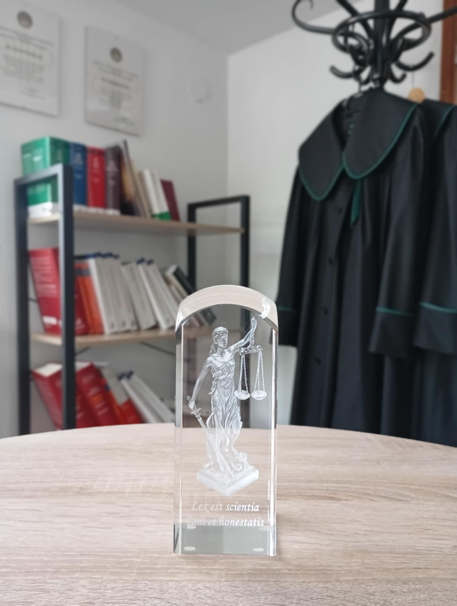 Kryształowa statuetka Temidy z łacińskim napisem, na drewnianym biurku, w tle togi adwokackie i regał z książkami, w kancelarii prawnej.