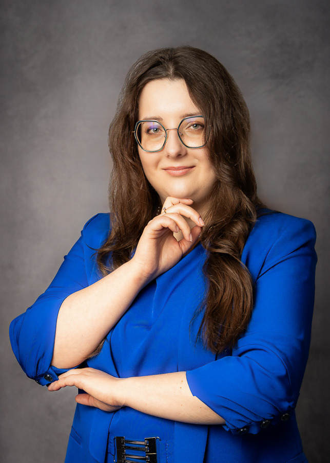 Portret kobiety w okularach i niebieskim żakiecie, z dłonią pod brodą, na szarym tle. Profesjonalny wizerunek.